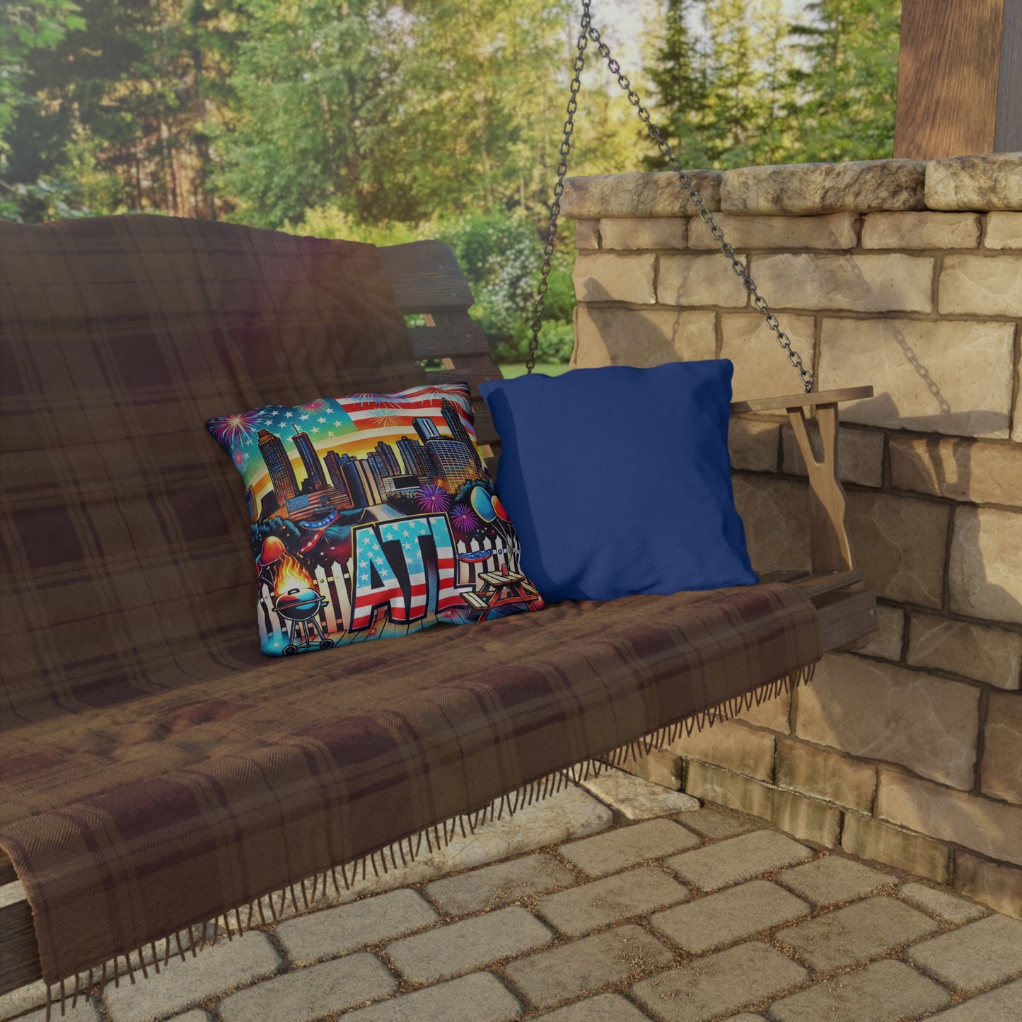Two throw pillows on an outdoor porch swing with a brown plaid blanket. One pillow features a colorful Atlanta skyline with fireworks, an American flag backdrop, and bold "ATL" lettering with BBQ and party details. The other pillow has a solid royal blue fabric back, providing a perfect contrast for outdoor or patio seating décor.