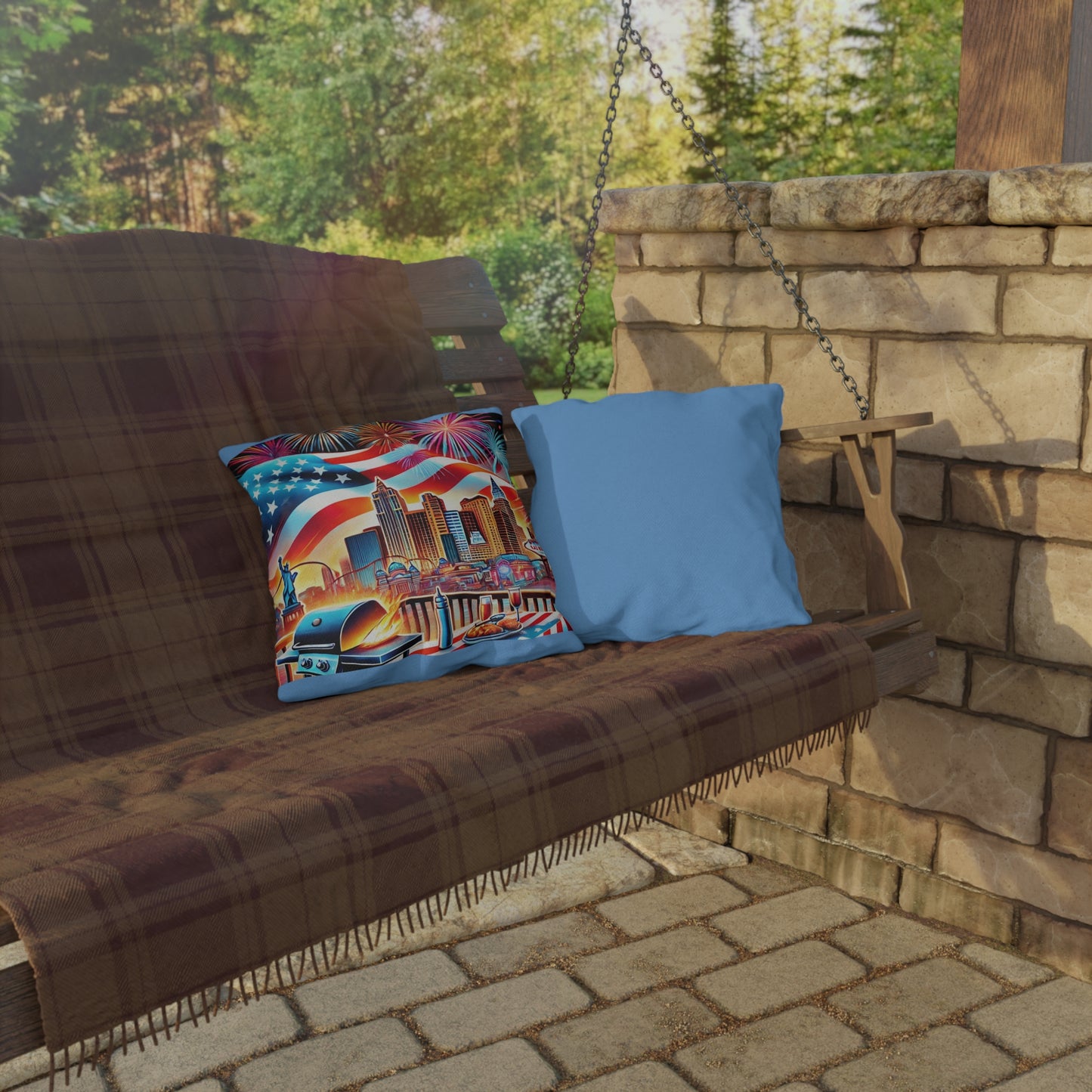 Two throw pillows on an outdoor swing bench with a brown plaid blanket. One pillow features a colorful Las Vegas skyline with fireworks, an American flag, and a BBQ scene, while the other pillow has a solid sky blue fabric back. The setup is ideal for outdoor gatherings, summer BBQs, or patriotic holiday décor.