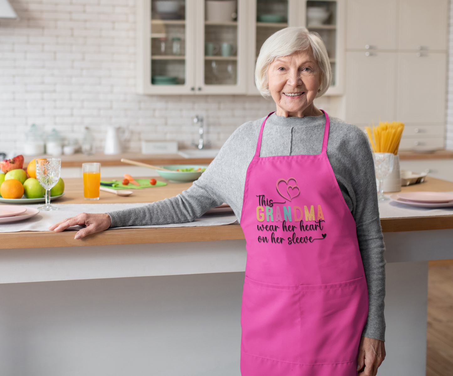 Elderly woman smiling in a bright modern kitchen while wearing a hot pink apron that says “This Grandma wears her heart on her sleeve” with heart graphics. A perfect promotional image for personalized gifts for grandmothers, ideal for birthdays, Mother’s Day, or holiday gifting.