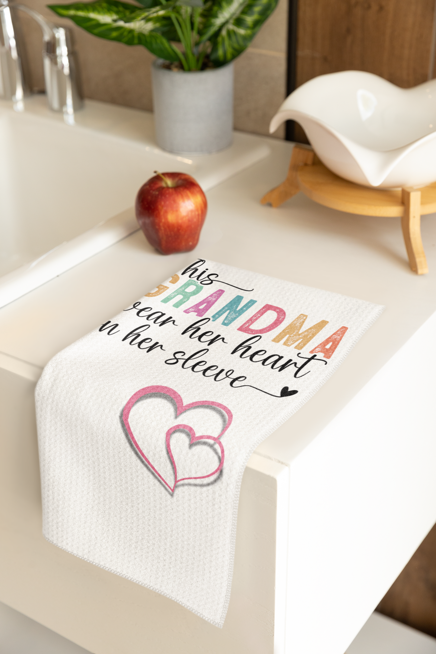 White waffle weave kitchen towel with colorful text reading 'This Grandma wears her heart on her sleeve' and two pink hearts, draped over a white countertop near a red apple, decorative bowl, and potted plant – perfect gift for grandmothers