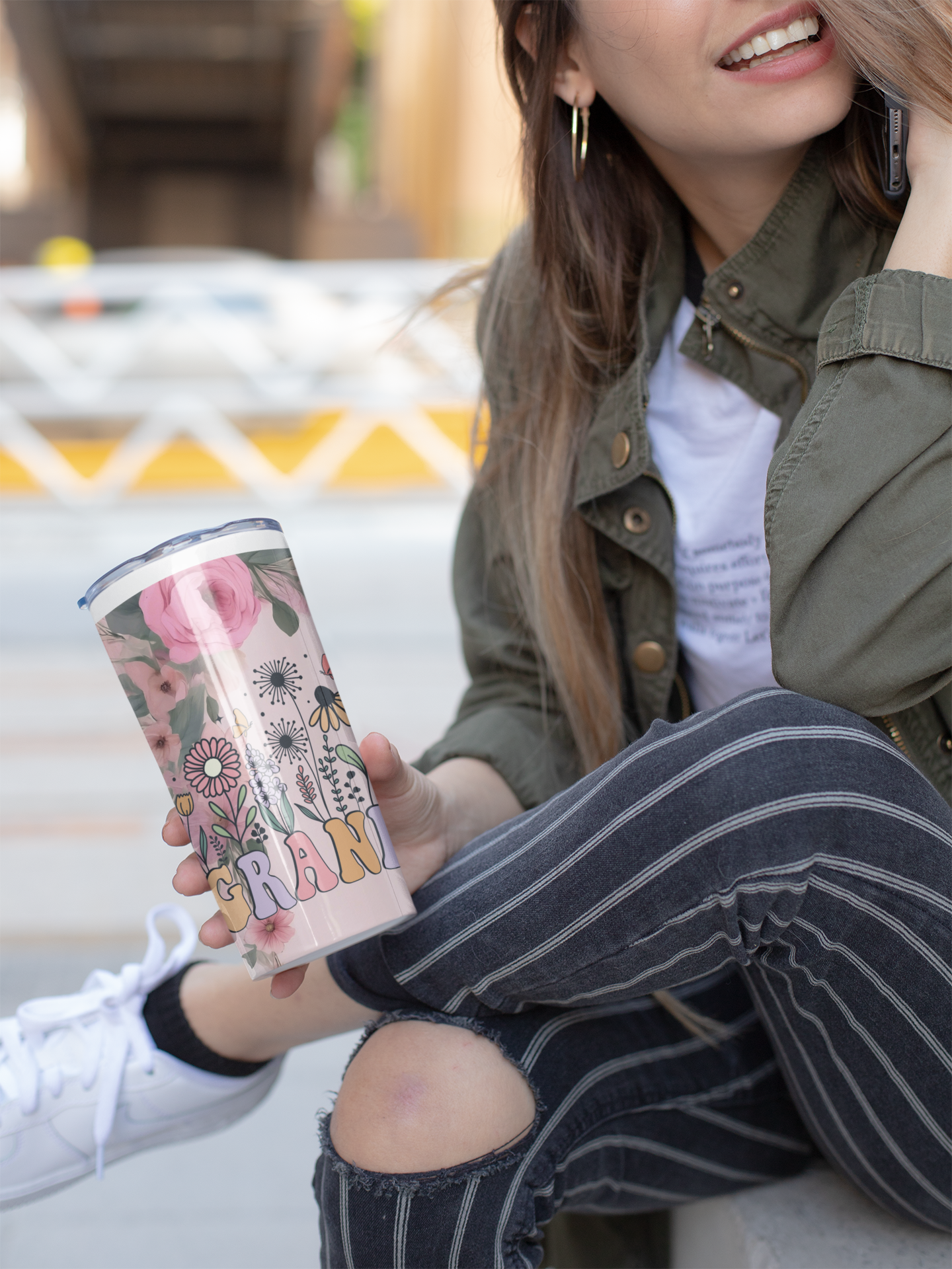 Stylish young woman sitting outdoors holding a pink floral "Grandma" tumbler with retro lettering and flower illustrations. Perfect lifestyle photo for promoting insulated drinkware as a thoughtful gift for grandmothers, Mother’s Day, or special celebrations.