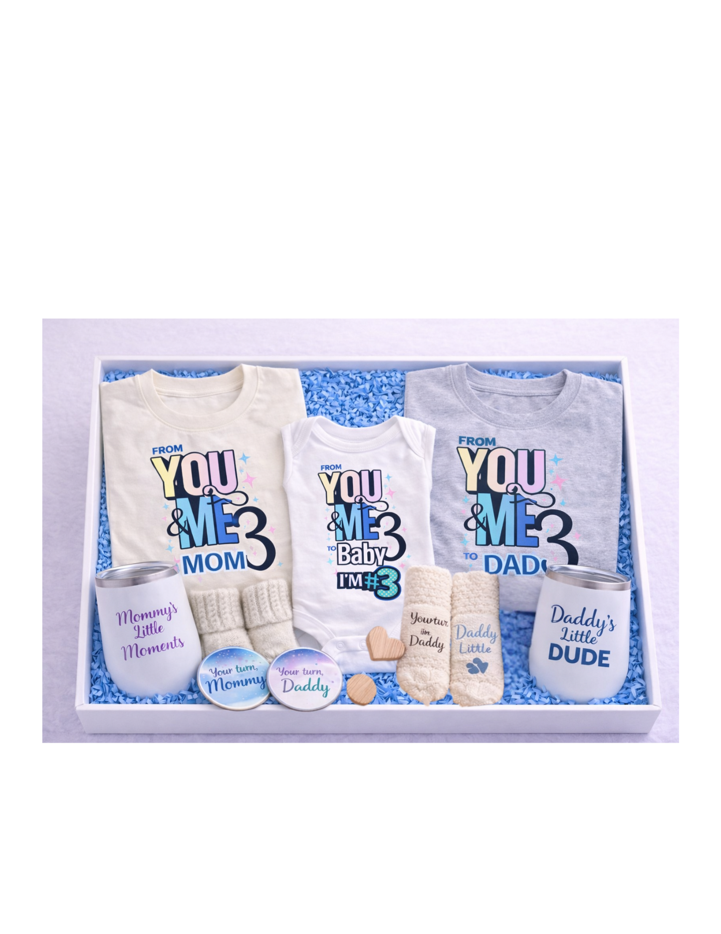 A curated family gift box in a white box with blue crinkle paper featuring matching “From You & Me to Us Three” shirts for mom and dad, a baby onesie, mommy and daddy wine tumblers, soft knit socks for mom, dad, and baby, and coordinating “Your turn Mommy” and “Your turn Daddy” coasters—perfect for baby announcements, showers, and new parent gifts.