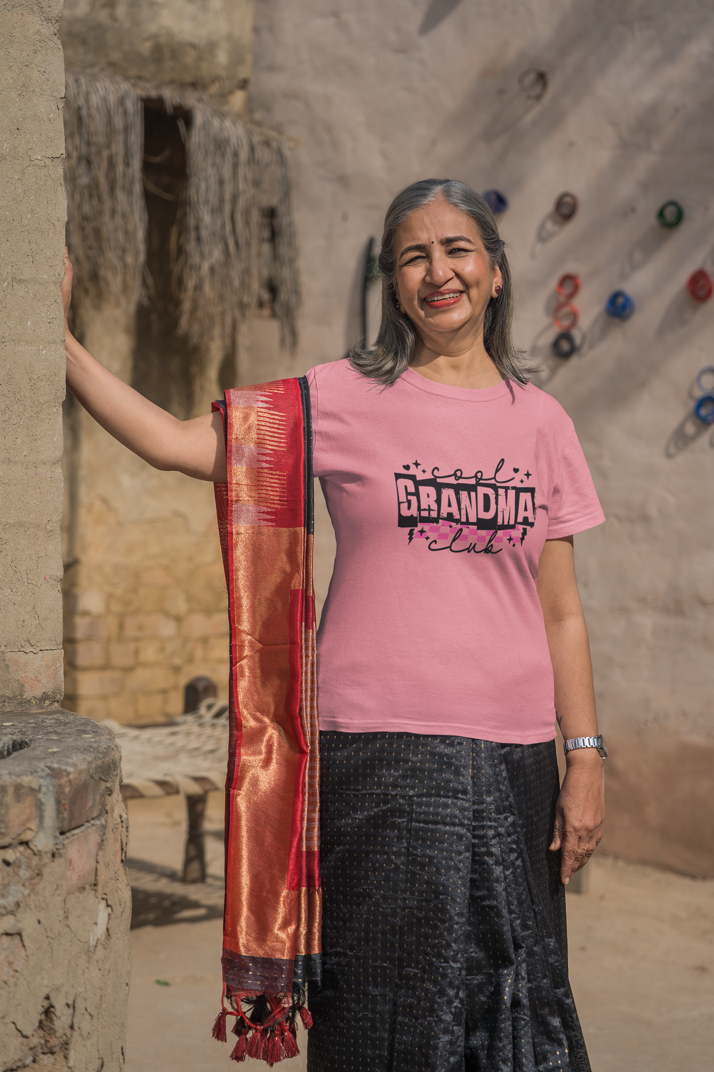 Confident older woman wearing a pink “Cool Grandma Club” T-shirt with bold black lettering, standing outdoors in traditional attire with a red and gold dupatta. Ideal lifestyle photo for showcasing unique and empowering gifts for grandmothers. Perfect for Mother’s Day, birthdays, or family celebrations.
