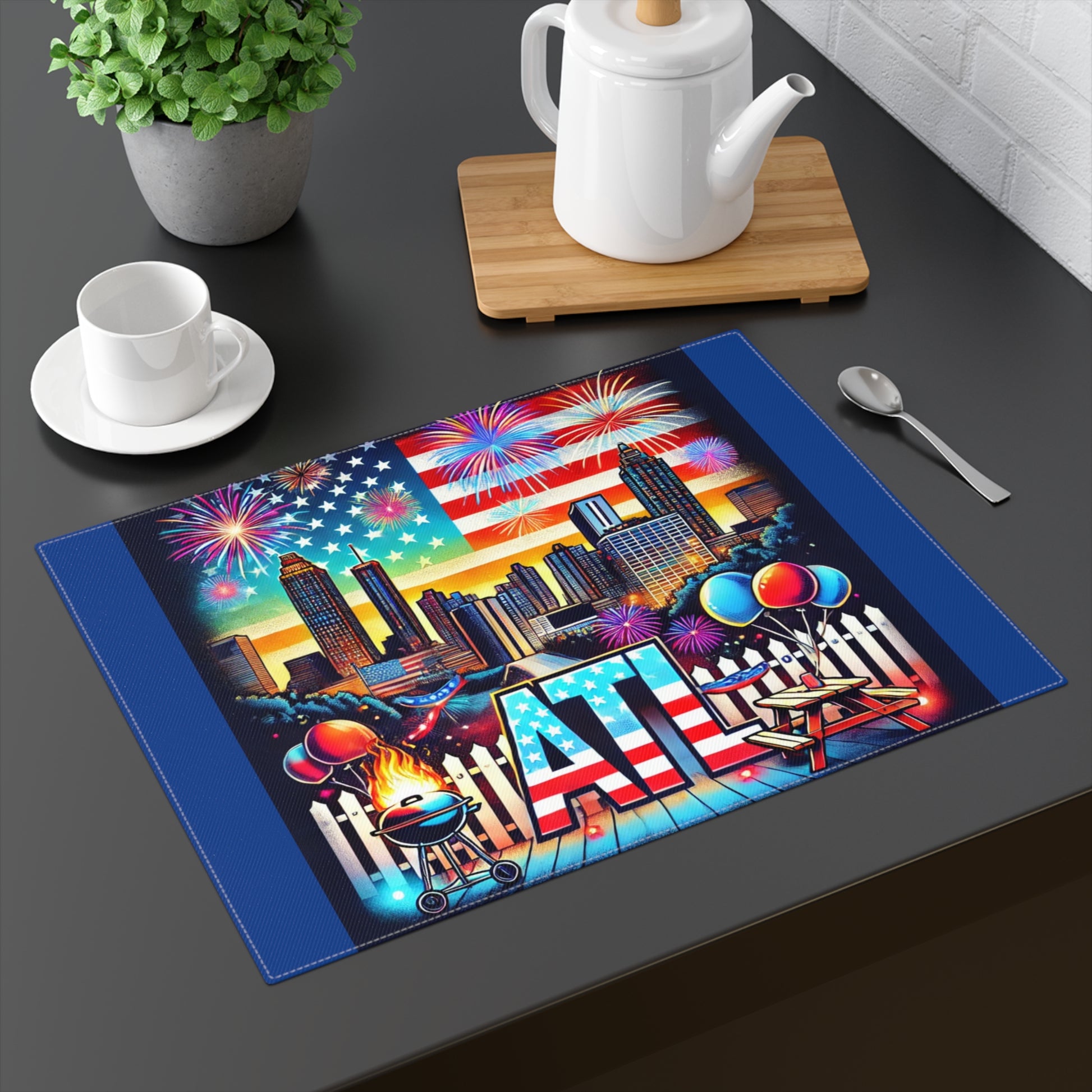 A vibrant Atlanta-themed placemat displayed on a black dining table with a teapot, cup, and spoon. The design features the Atlanta skyline with tall buildings, a large American flag, and colorful fireworks in the sky. The foreground includes BBQ grill flames, picnic tables, balloons, and bold "ATL" lettering with a stars and stripes pattern, making it perfect for 4th of July, summer parties, and Atlanta-themed décor.