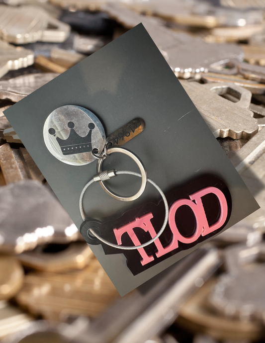  A stylish TLOD keychain with a black and pink acrylic design, attached to a silver keyring featuring a round metal charm with a crown engraving and a small rectangular tag with "1964" inscribed. The keychain is placed on a black background and overlaid onto an image of scattered silver keys.
