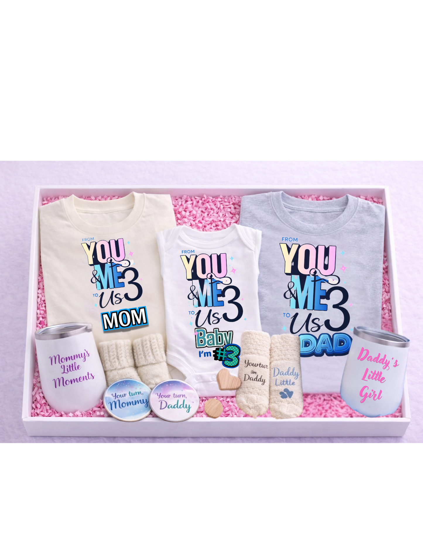 A premium family gift box arranged in a white box with pink crinkle paper, featuring matching “From You & Me to Us Three” shirts for mom, dad, and baby, a baby onesie, mommy and daddy wine tumblers, cozy socks for each family member, and coordinating “Your turn Mommy” and “Your turn Daddy” coasters—perfect for baby announcements, showers, or new parent gifts.