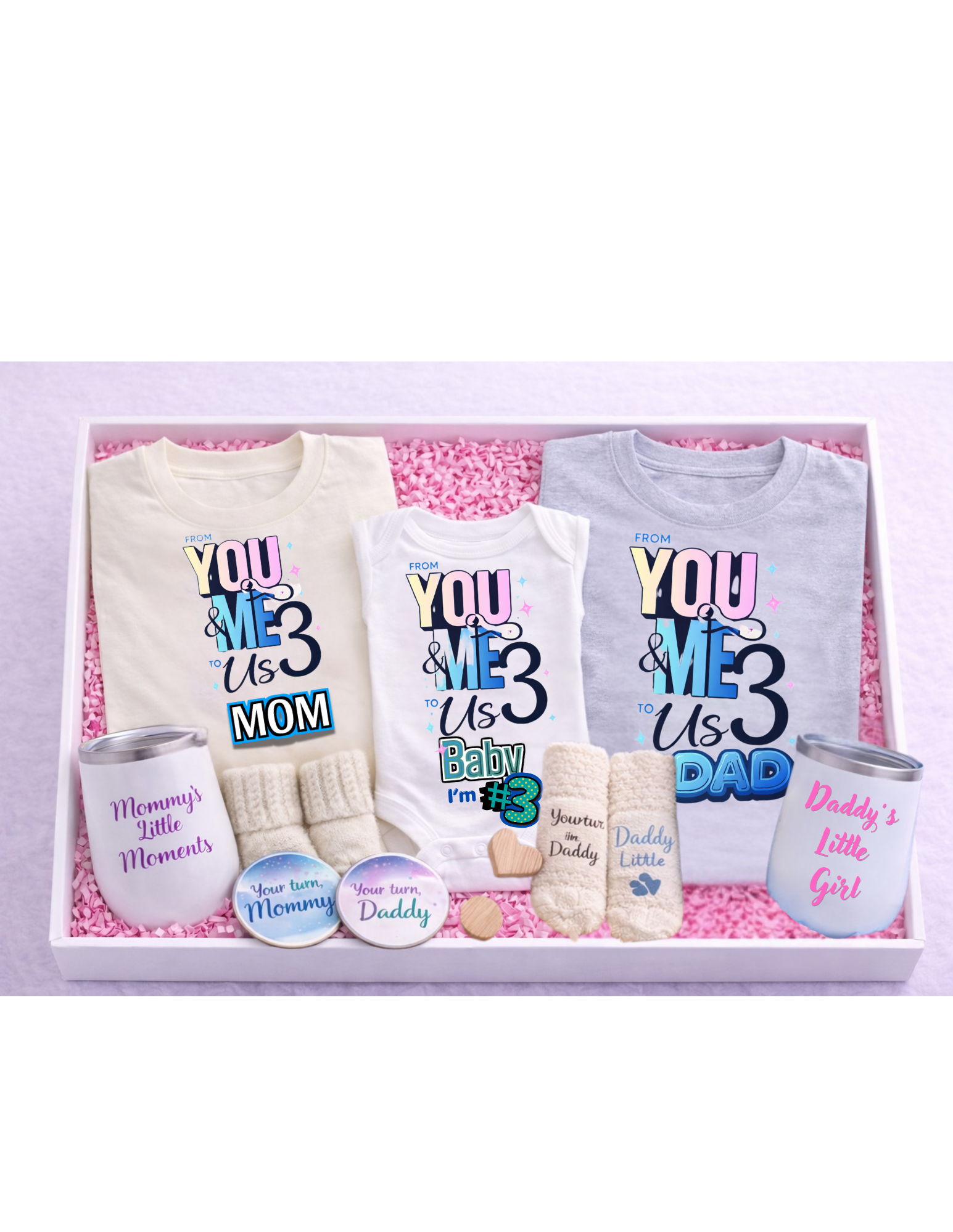 A premium family gift box arranged in a white box with pink crinkle paper, featuring matching “From You & Me to Us Three” shirts for mom, dad, and baby, a baby onesie, mommy and daddy wine tumblers, cozy socks for each family member, and coordinating “Your turn Mommy” and “Your turn Daddy” coasters—perfect for baby announcements, showers, or new parent gifts.