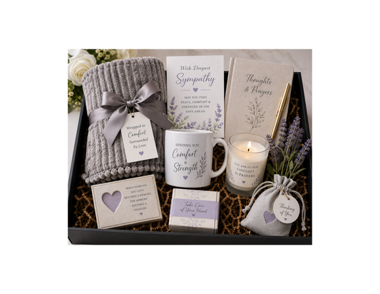 A sympathy and comfort gift basket arranged in a black tray, featuring a folded gray knit blanket tied with a gray ribbon, a white mug reading 'Sending You Comfort & Strength,' a candle, cards, a small box, a lavender sachet, and a notebook on a light marble surface.