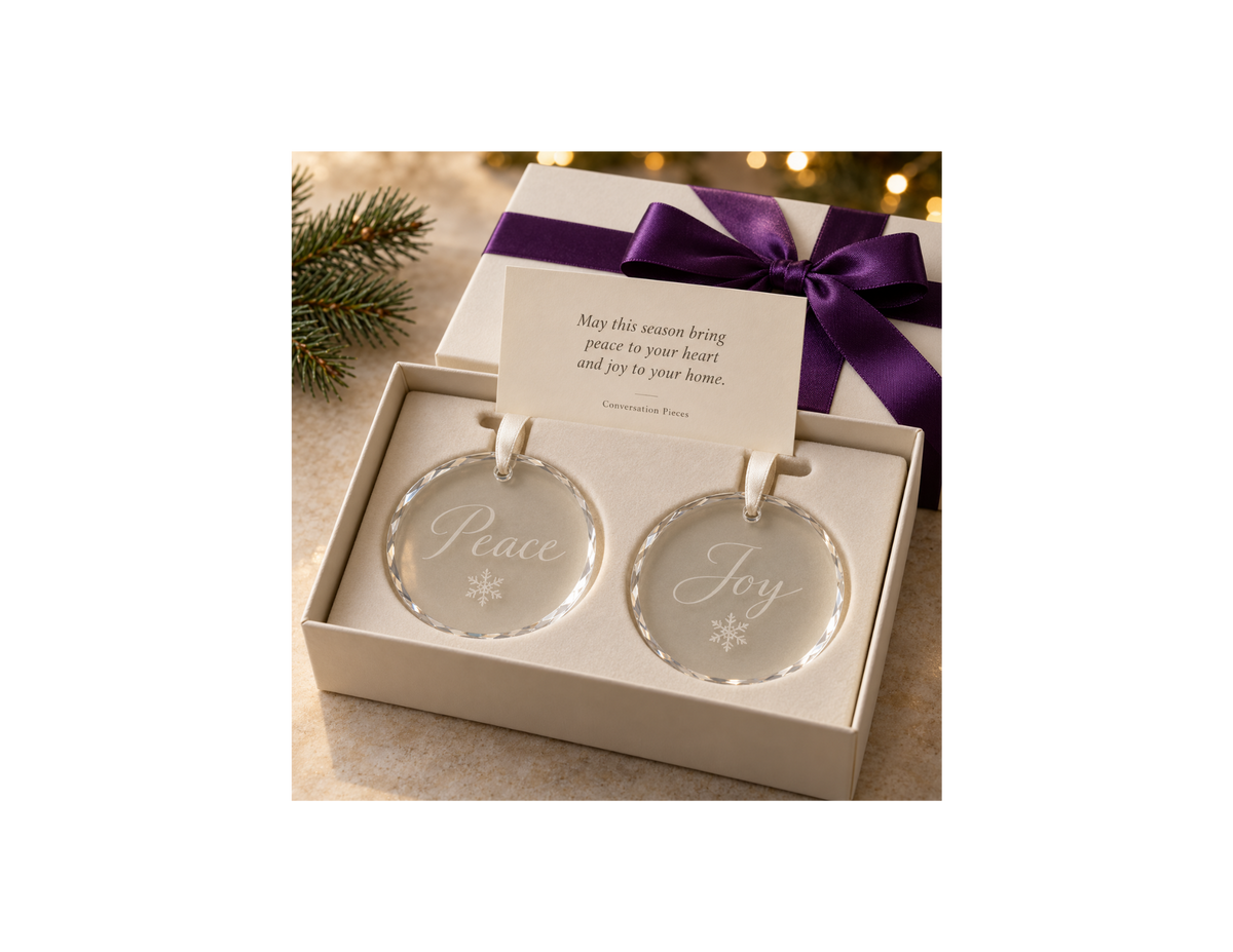 Two round crystal Christmas ornaments engraved with Peace, Joy, and snowflakes, with ivory hanging ribbons in an ivory gift box with purple ribbon and a holiday keepsake card.