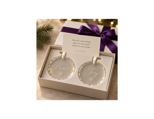 Two round crystal Christmas ornaments engraved with Peace, Joy, and snowflakes, with ivory hanging ribbons in an ivory gift box with purple ribbon and a holiday keepsake card.