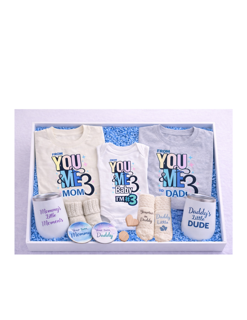A curated family gift box in a white box with blue crinkle paper featuring matching “From You & Me to Us Three” shirts for mom and dad, a baby onesie, mommy and daddy wine tumblers, soft knit socks for mom, dad, and baby, and coordinating “Your turn Mommy” and “Your turn Daddy” coasters—perfect for baby announcements, showers, and new parent gifts.
