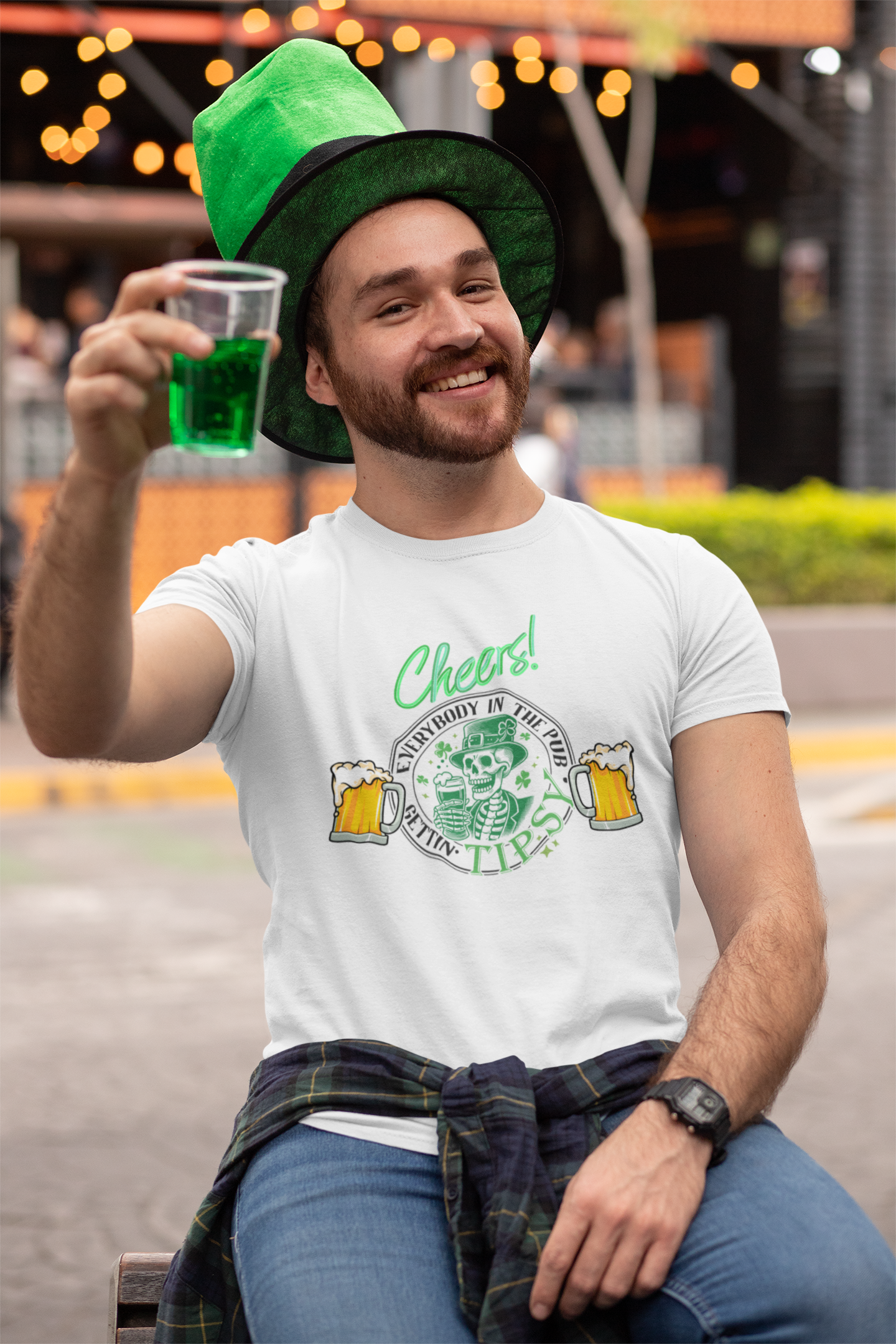 A cheerful man wearing a festive white St. Patrick’s Day t-shirt with a "Cheers!" design featuring a skeleton holding a beer mug, surrounded by the phrase "Everybody in the Pub Gettin’ Tipsy" and two frothy beer mugs. He is sitting outdoors at a celebration, dressed in a green leprechaun-style hat and a plaid shirt tied around his waist. He raises a cup of green beer with a big smile, embracing the lively St. Paddy’s Day atmosphere.